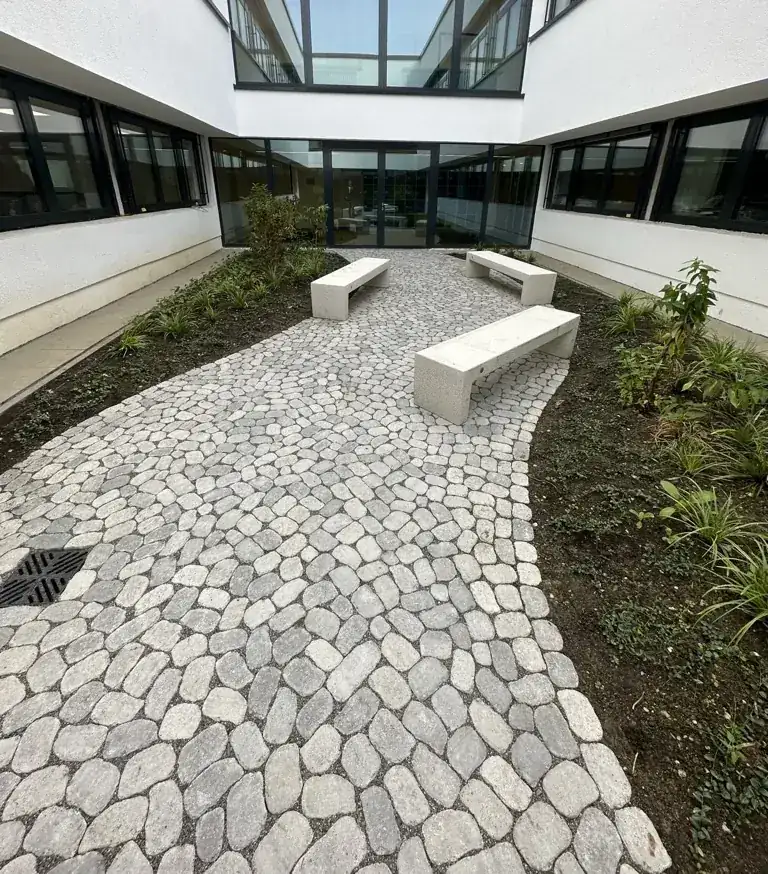Geschwungener Weg aus Betonpflaster ARENA mit runden Kanten in einem Innenhof, daneben Beete und weiße Bänke