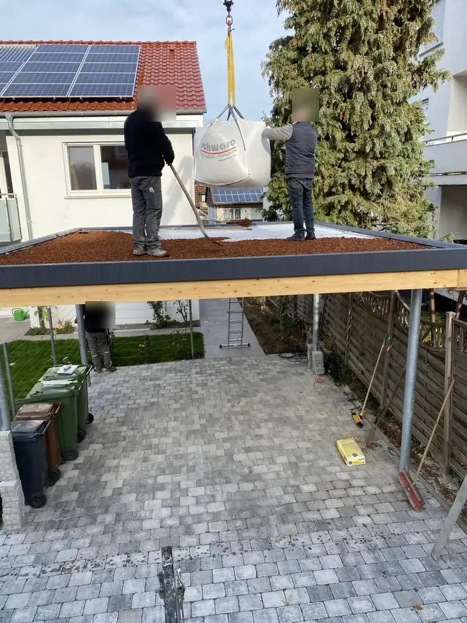 Gründach auf einem Carport im Bau: Ein Kran hebt einen Big Bag mit Dachsubstrat aufs Dach, zwei Mitarbeiter verteilen das Substrat