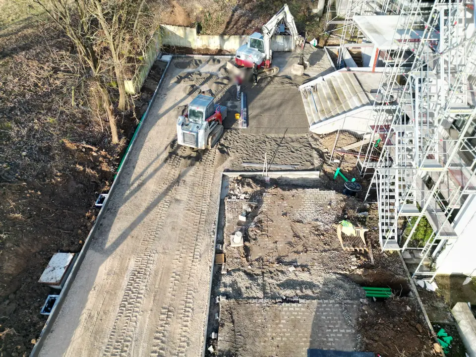 Großbaustelle an einem Firmengebäude von oben: Bagger und Kompaktlader auf der verdichteten Tragschicht, gesetzte Randsteine, neue Entwässerungsleitungen