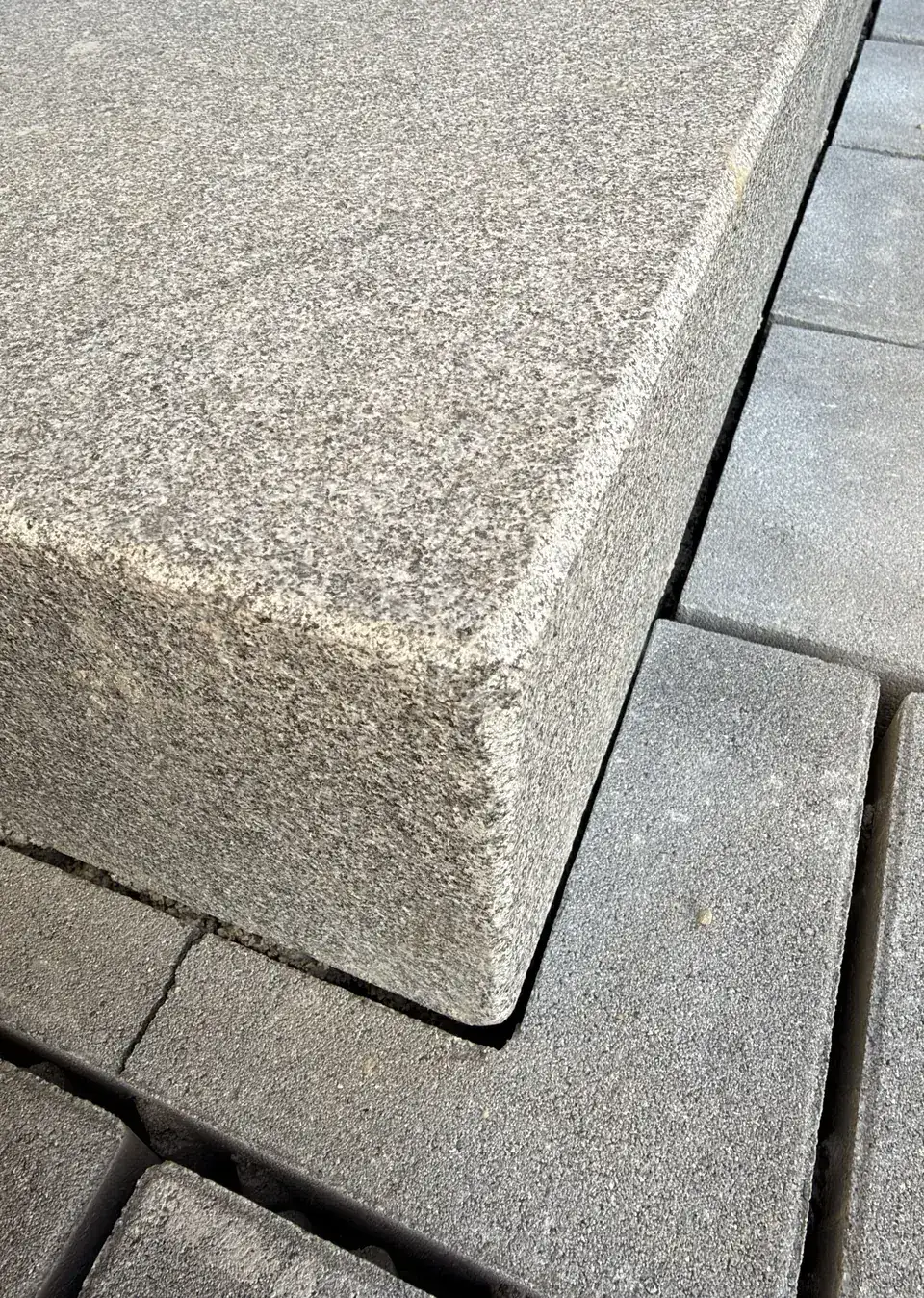 Ecke einer Blockstufe aus anthrazitfarbenem Granit mit fein gebrochener Kante über dem Betonpflaster