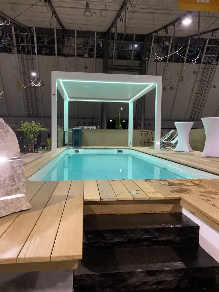 Pool mit Holzdeck, Natursteintreppe und weißer Pergola mit Lichtleiste auf dem Messestand 2019