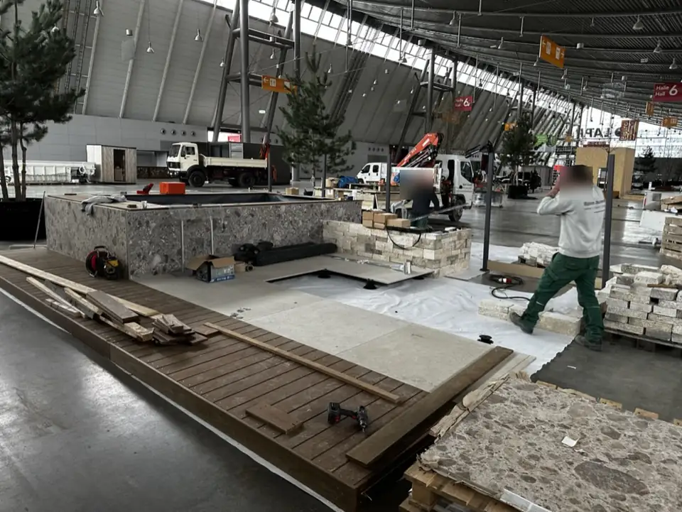 Aufbau des Messestands: Holzdeck, Natursteinverkleidung am Becken, Mauersteine auf Paletten, im Hintergrund LKW mit Kran