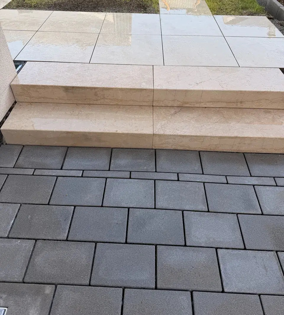 Zwei Natursteinstufen zwischen Betonpflaster und Terrasse aus Keramikplatten, dahinter Trittplatten im Rasen