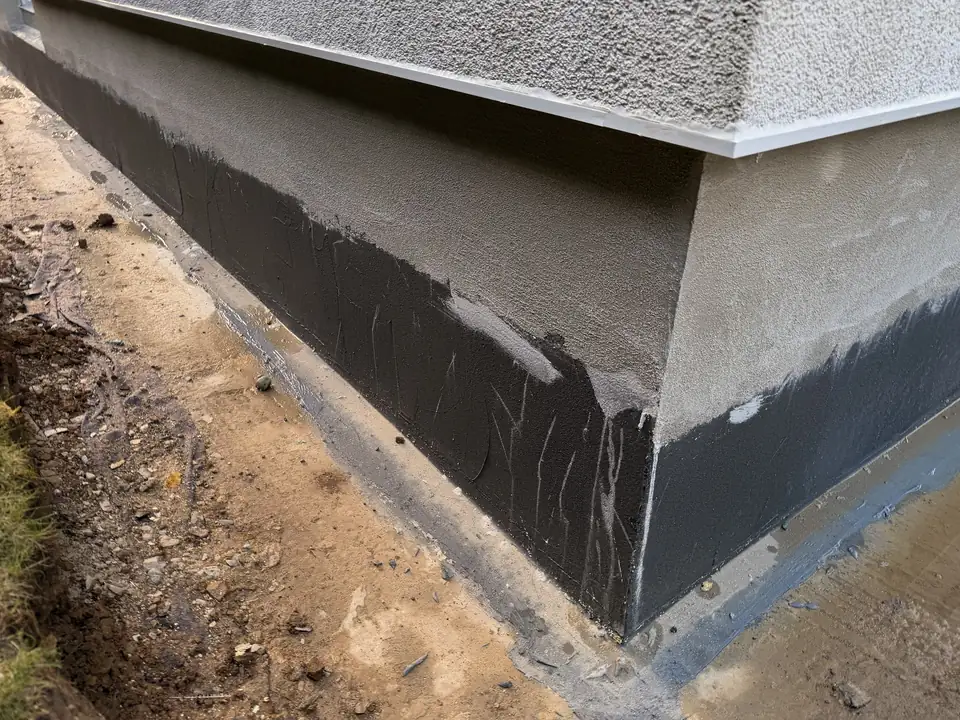 Gebäudeecke mit neuer dunkler Sockelabdichtung, darüber frischer Sockelputz und Fassade, davor der freigelegte Graben