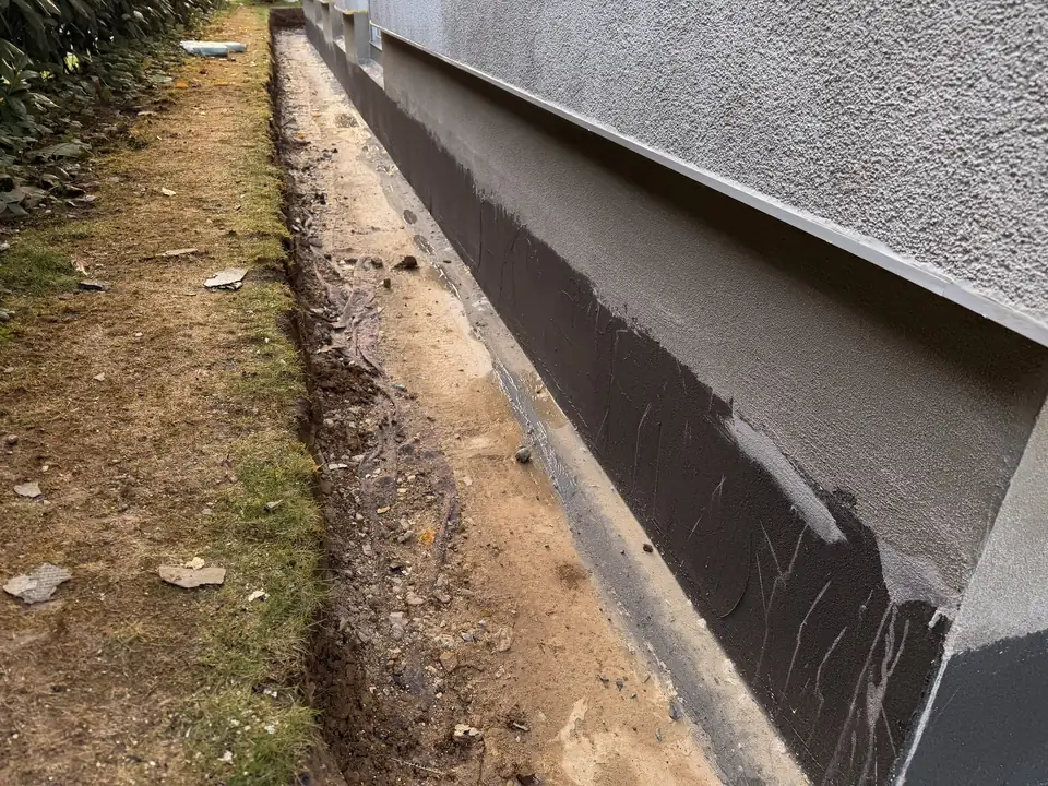 Lange Hauswand mit neuer dunkler Sockelabdichtung und frischem Sockelputz, daneben der offene Graben