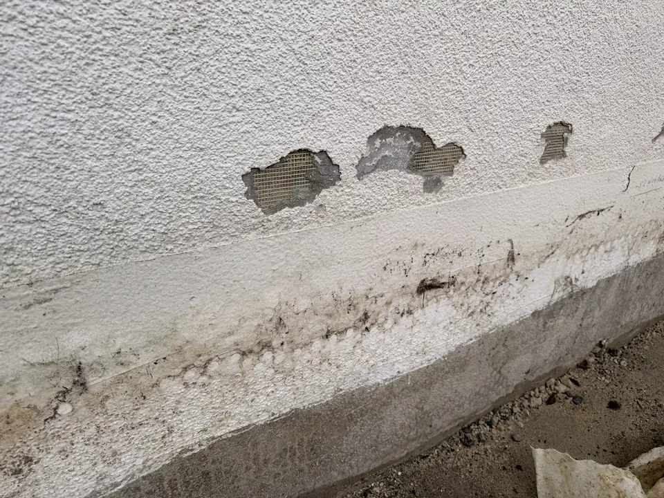 Nahaufnahme: Putzschäden mit sichtbarem Armierungsgewebe und Wasserrändern am Sockel