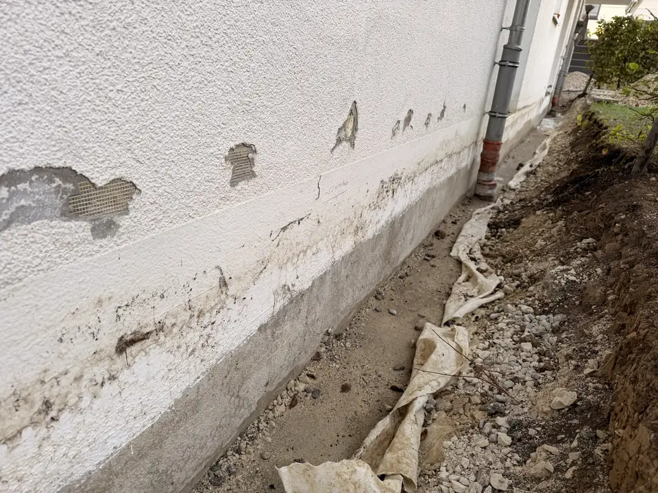 Hauswand mit abgeplatztem Putz und freiliegendem Gewebe, darunter verschmutzter Sockel und freigelegter Graben